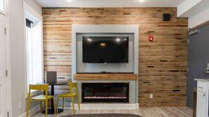 A modern living room with a flat-screen TV mounted above a fireplace, wooden panel wall, two yellow chairs, and a black ottoman. - Downtown+HotTub+Wifi+FreeParking @151 Spring Ste A (Eureka Springs)