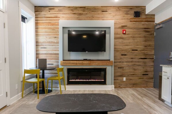 A modern living room with a flat-screen TV mounted above a fireplace, wooden panel wall, two yellow chairs, and a black ottoman. - Downtown+HotTub+Wifi+FreeParking @151 Spring Ste A (Eureka Springs)