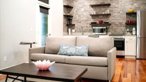 A modern kitchen with a grey brick backsplash, wooden shelves, a stainless steel refrigerator, and a beige sofa with a decorative pillow. - Downtown+HotTub+Wifi+FreeParking @151 Spring Ste F (Eureka Springs)