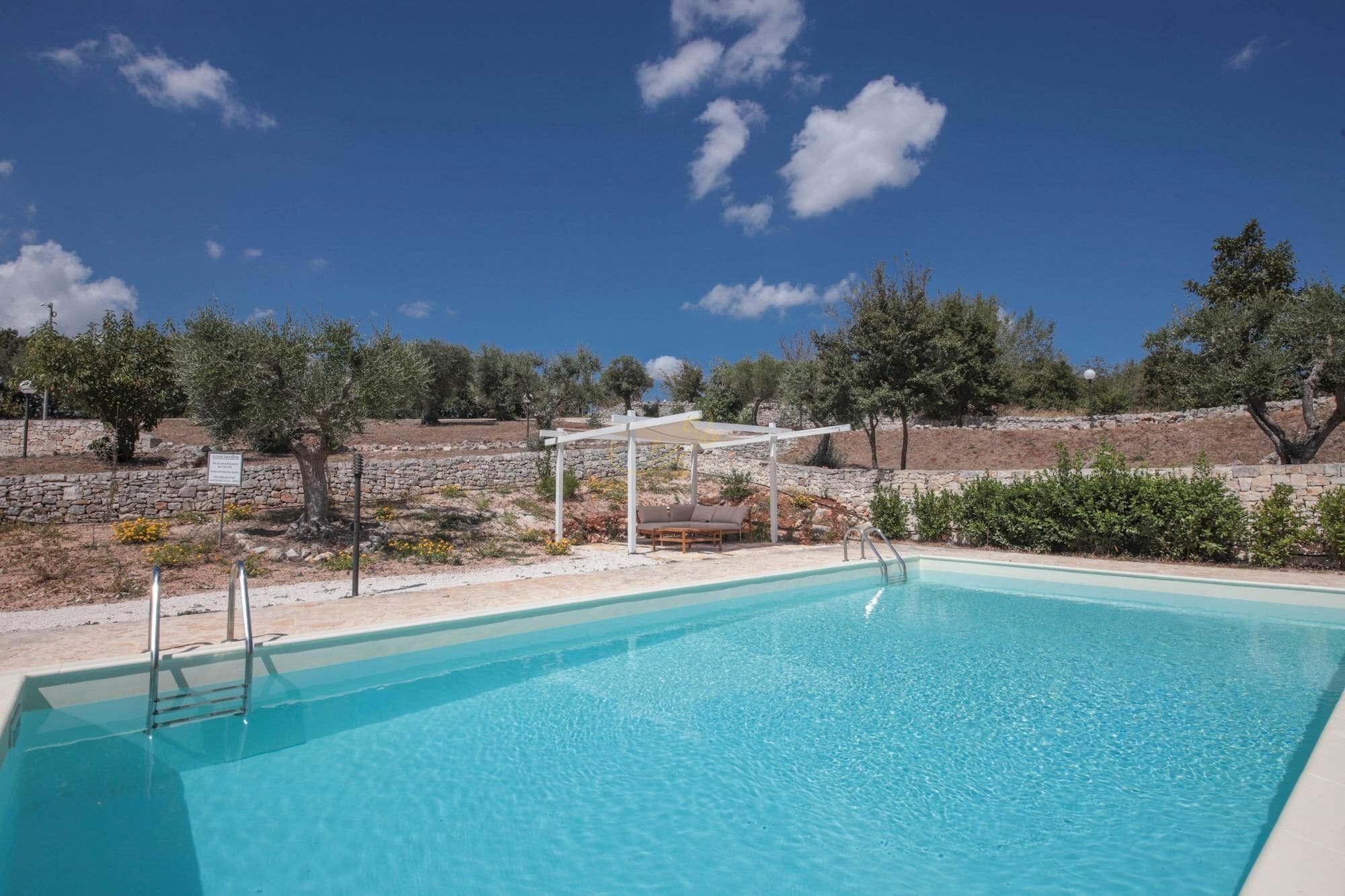 A clear blue pool with a stone wall and trees in the background.