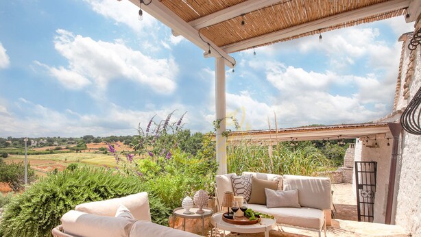 A covered outdoor seating area with wicker furniture, a table with a wine bottle, and a view of the countryside. - TD Trulli Altura With Pool Stunning Sunsets (Martina Franca)