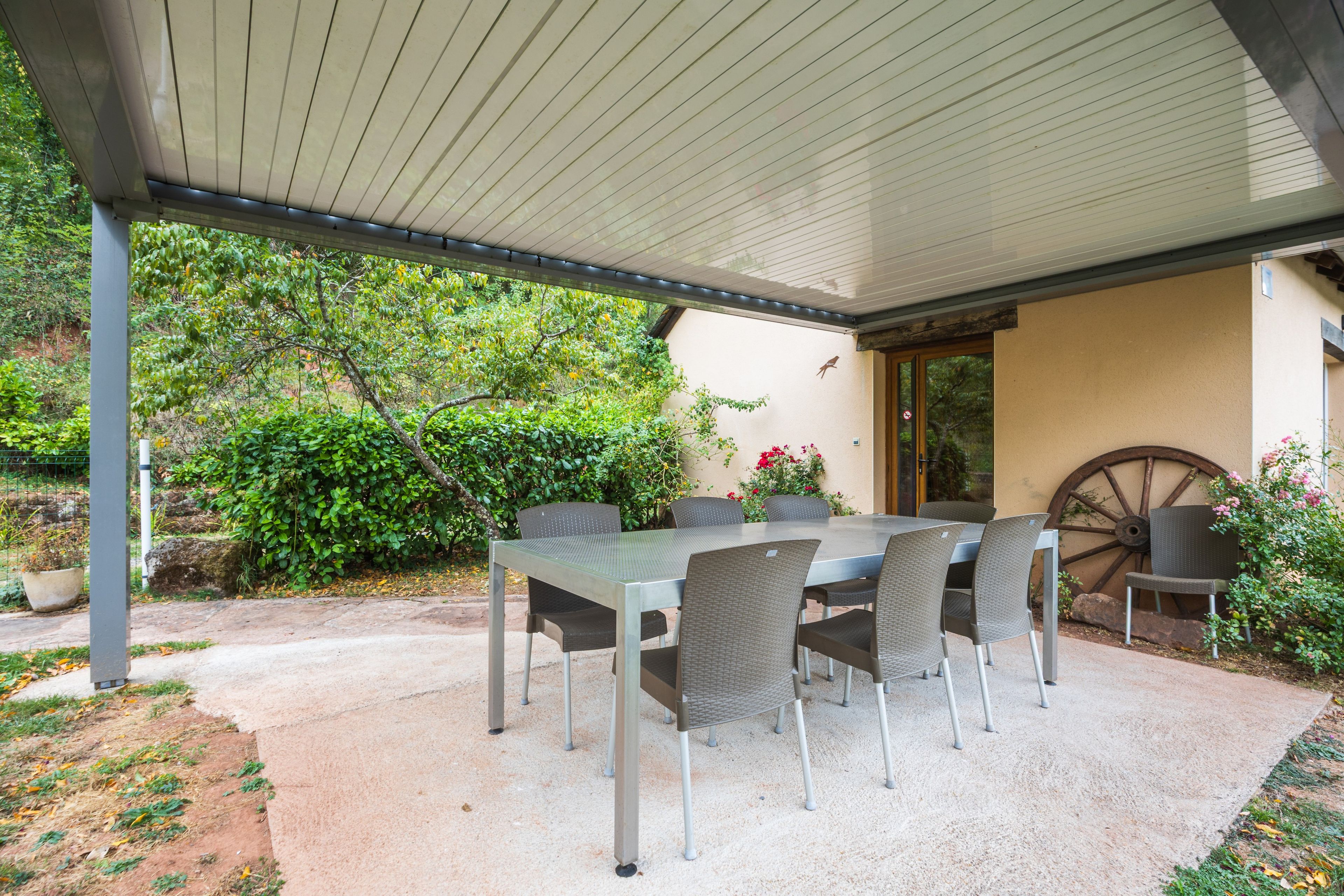 An outdoor patio with a covered table and chairs, surrounded by greenery and a decorative wheel.