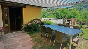 A covered outdoor dining area with a table and chairs, a wooden door, and a view of a green landscape.