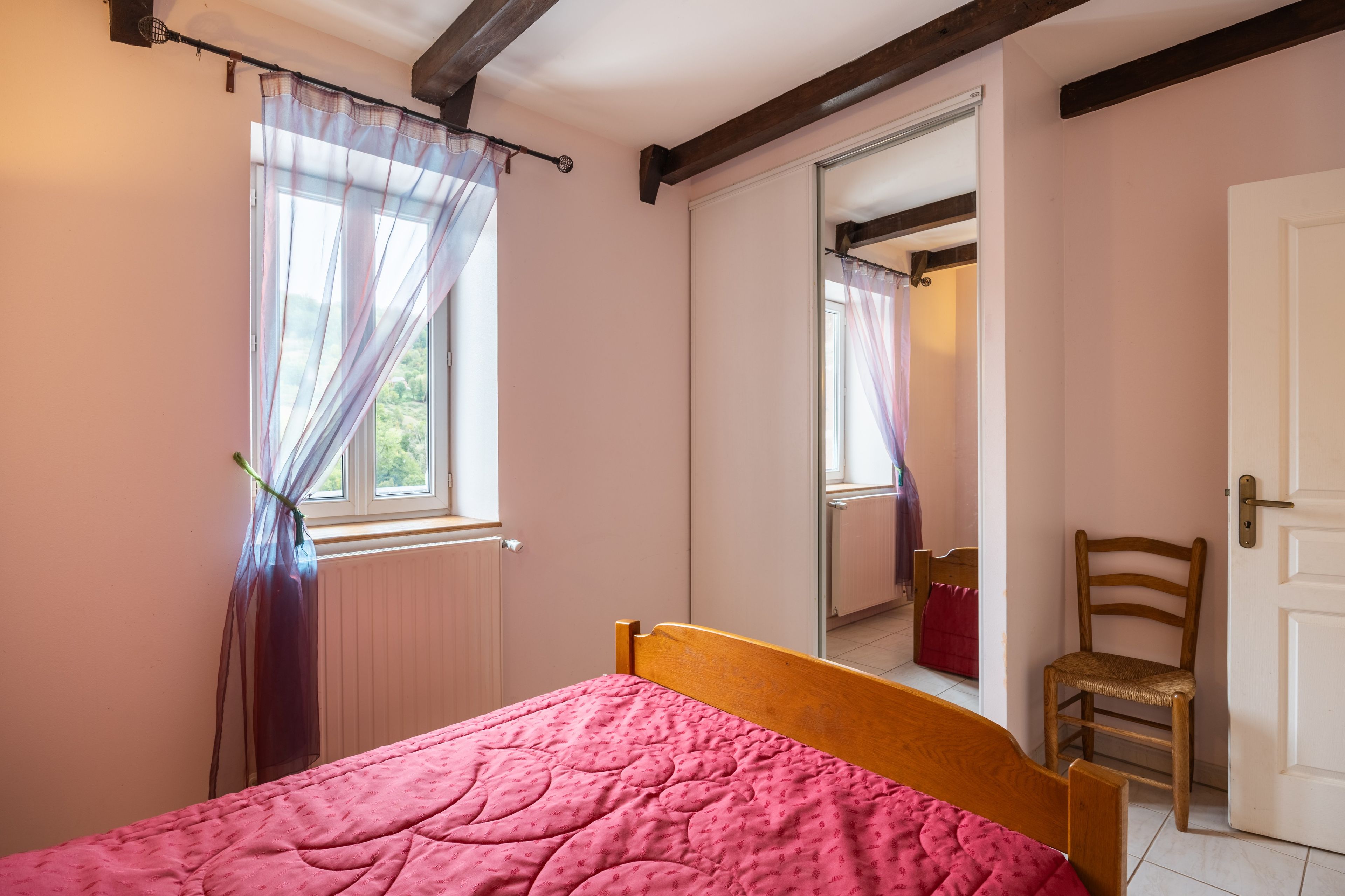 Un dormitorio con cama, una silla, un ventanal con cortinas y una puerta.