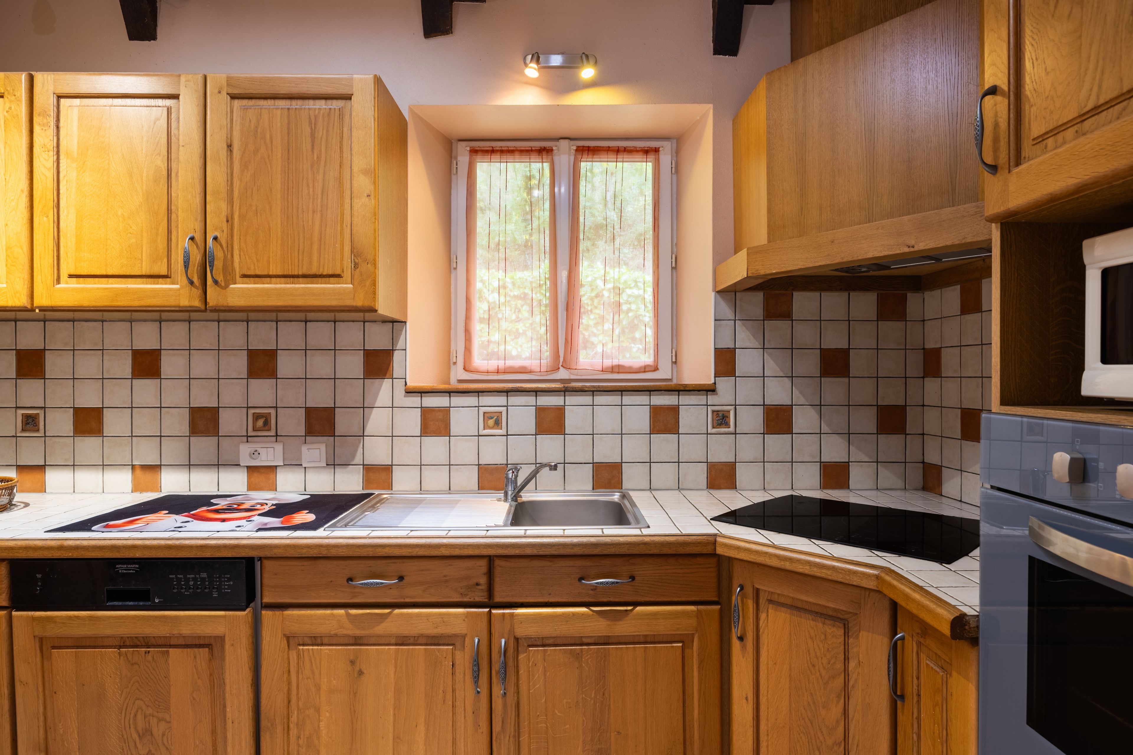 Une cuisine avec des meubles de rangement en bois, un pare-feu en carrelage, une fenêtre avec des rideaux et un four à micro-ondes.
