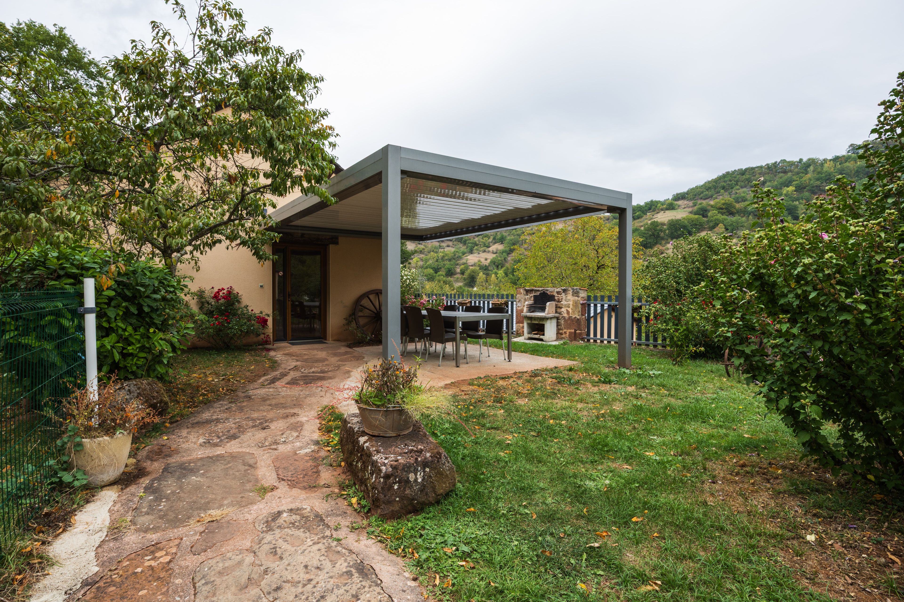 Un espace extérieur aménagé, abrité et doté de sièges, avec un chemin de pierre menant à un bâtiment, le tout entouré de verdure et de collines.