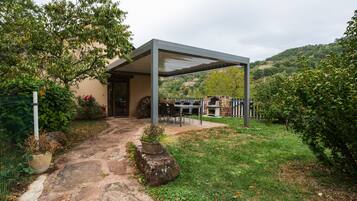 Un espace extérieur aménagé, abrité et doté de sièges, avec un chemin de pierre menant à un bâtiment, le tout entouré de verdure et de collines.