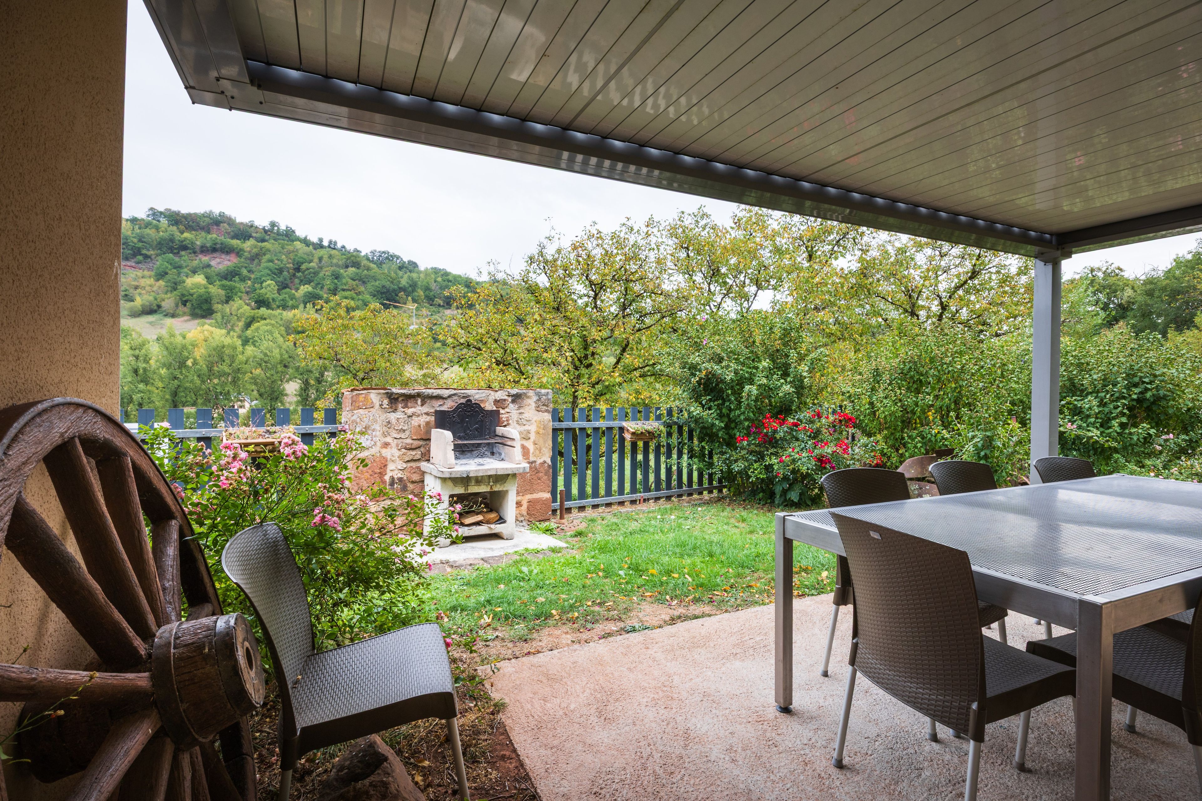 Um pátio externo com mesa e cadeiras, churrasqueira de pedra e vista para um jardim exuberante e colinas.