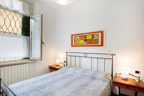 A bedroom with a bed, a bedside table with a lamp, a wall-mounted artwork, and a window with a metal screen.