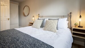 Cama bem arrumada com edredom branco, manta cinza e branca estampada e várias almofadas. Há duas mesas de cabeceira, uma com luminária e um pequeno relógio. Uma porta de madeira é visível à esquerda.