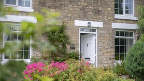 Un edificio de piedra con puerta y ventanas blancas, rodeado de exuberante vegetación y flores rosadas.