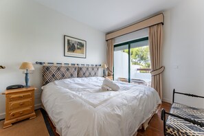 Una cama bien hecha con edredón blanco, una mesita de noche de madera con lámpara, cabecero estampado y una ventana con cortinas.