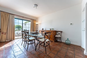 Uma sala de jantar com mesa de vidro, cadeiras de madeira, um armário de madeira e uma grande janela com cortinas.