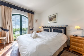 Uma cama bem arrumada com uma toalha, uma mesa de cabeceira de madeira com uma luminária, uma cadeira e uma janela grande com cortinas.