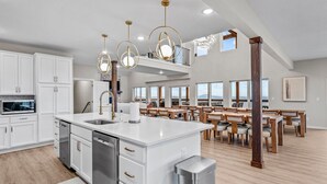 Una cocina amplia con muebles blancos, electrodomésticos de acero inoxidable y una isla grande. Hay lámparas colgantes sobre la isla y un comedor con mesas y sillas de madera.