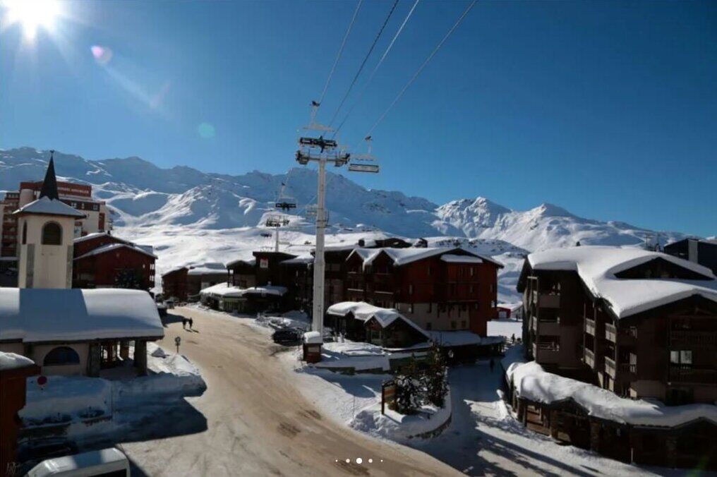 A snow-covered village with traditional wooden buildings, a church, and a ski lift against a backdrop of mountains.