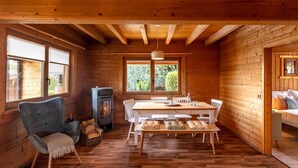Interior acogedor de una cabaña con una mesa de comedor de madera, sillas blancas, una chimenea y ventanas amplias con vistas al follaje.