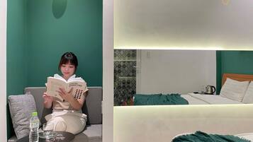 Una persona leyendo un libro en una habitación con pared verde, una cama y una mesita con una botella y vasos.