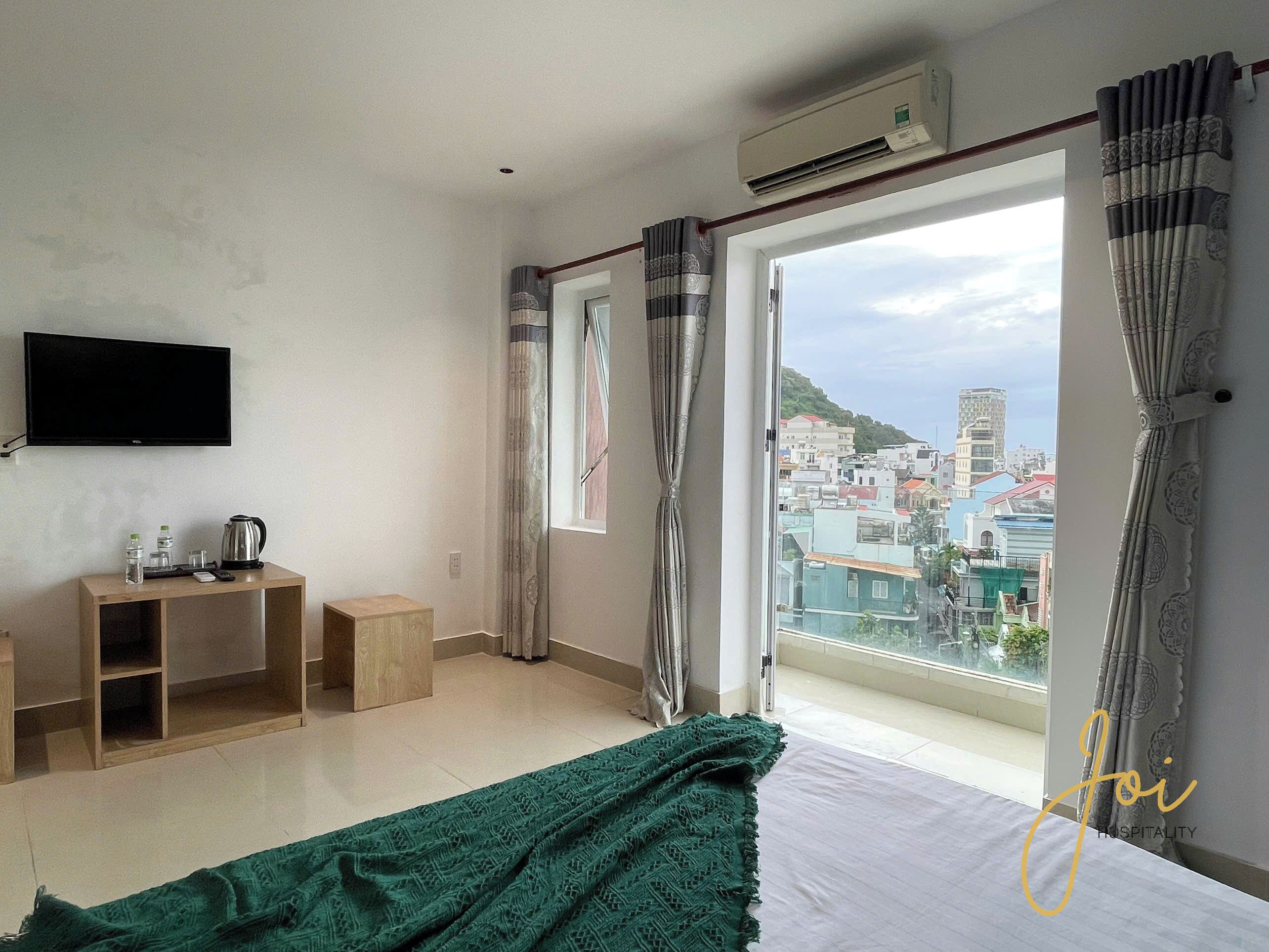 Une chambre d’hôtel avec un lit, une télévision, une petite table en bois avec une bouilloire, et une vue sur le paysage urbain à travers de grandes fenêtres.