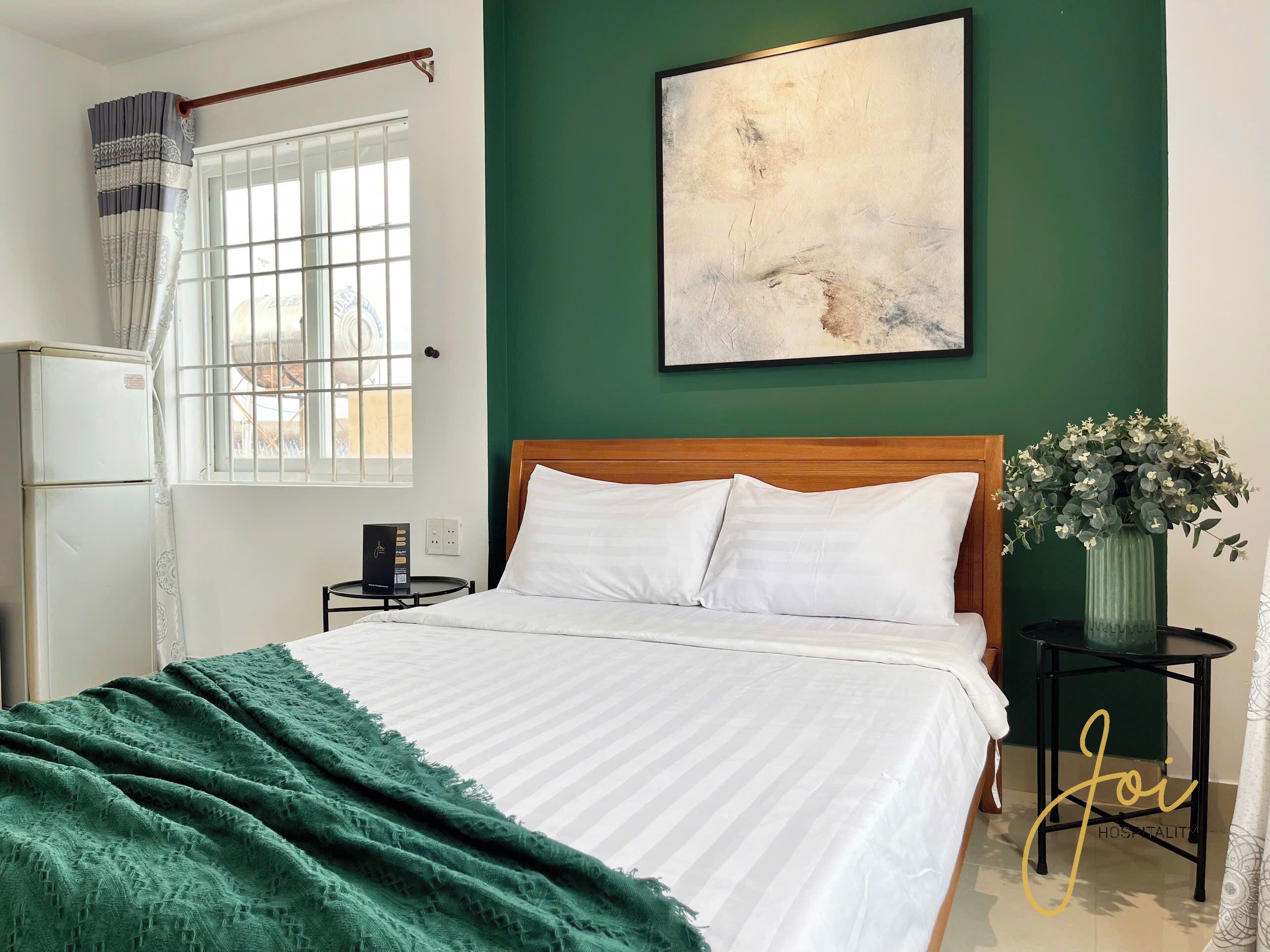 A bedroom with a bed, a green wall, a window with curtains, a painting, and a vase with flowers.