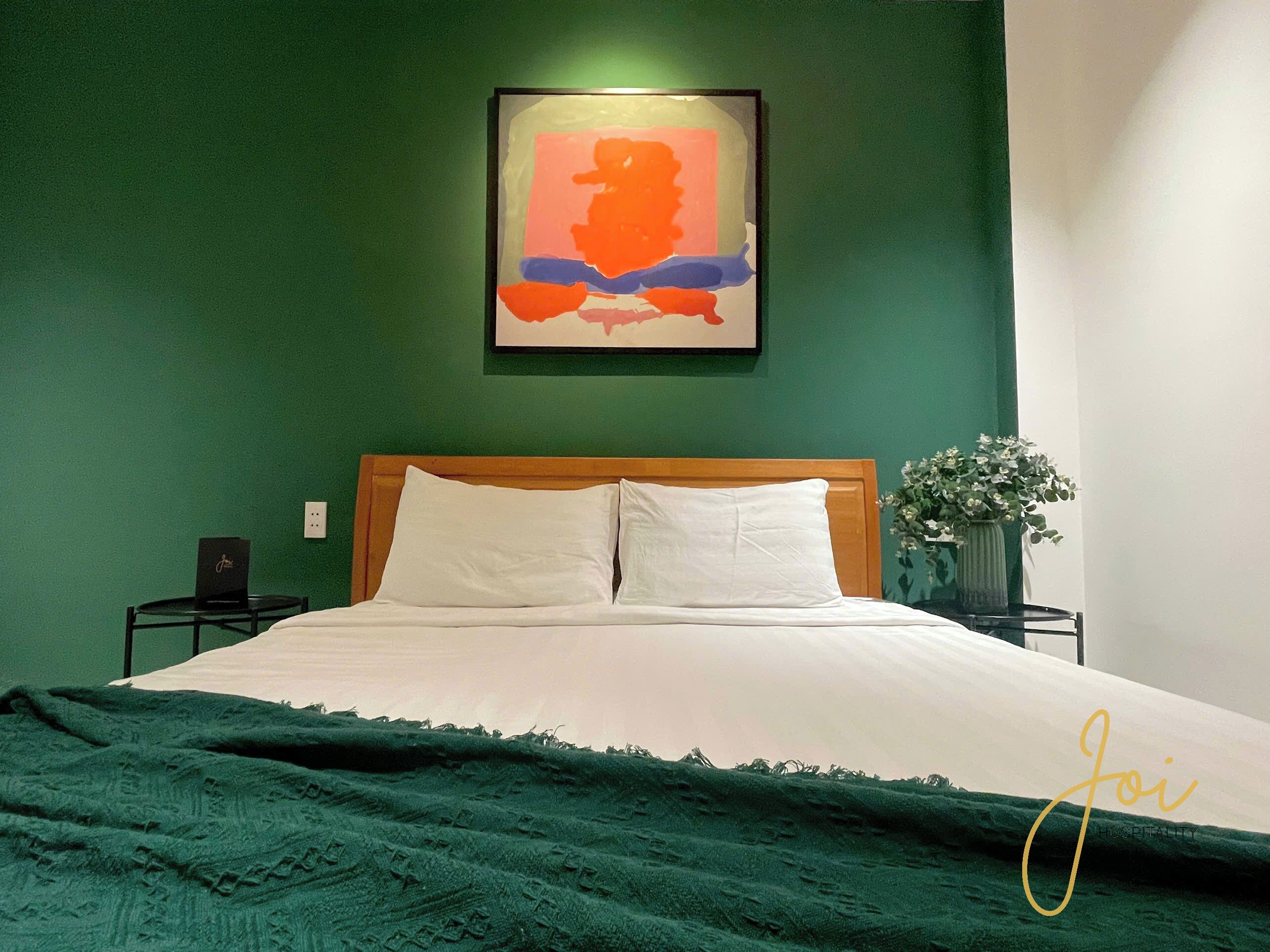 A neatly made bed with white linens and a green blanket, a framed abstract painting above it, and a small table with a lamp beside the bed.