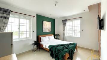A bedroom with a bed, a nightstand, a window with curtains, and a wall-mounted TV.