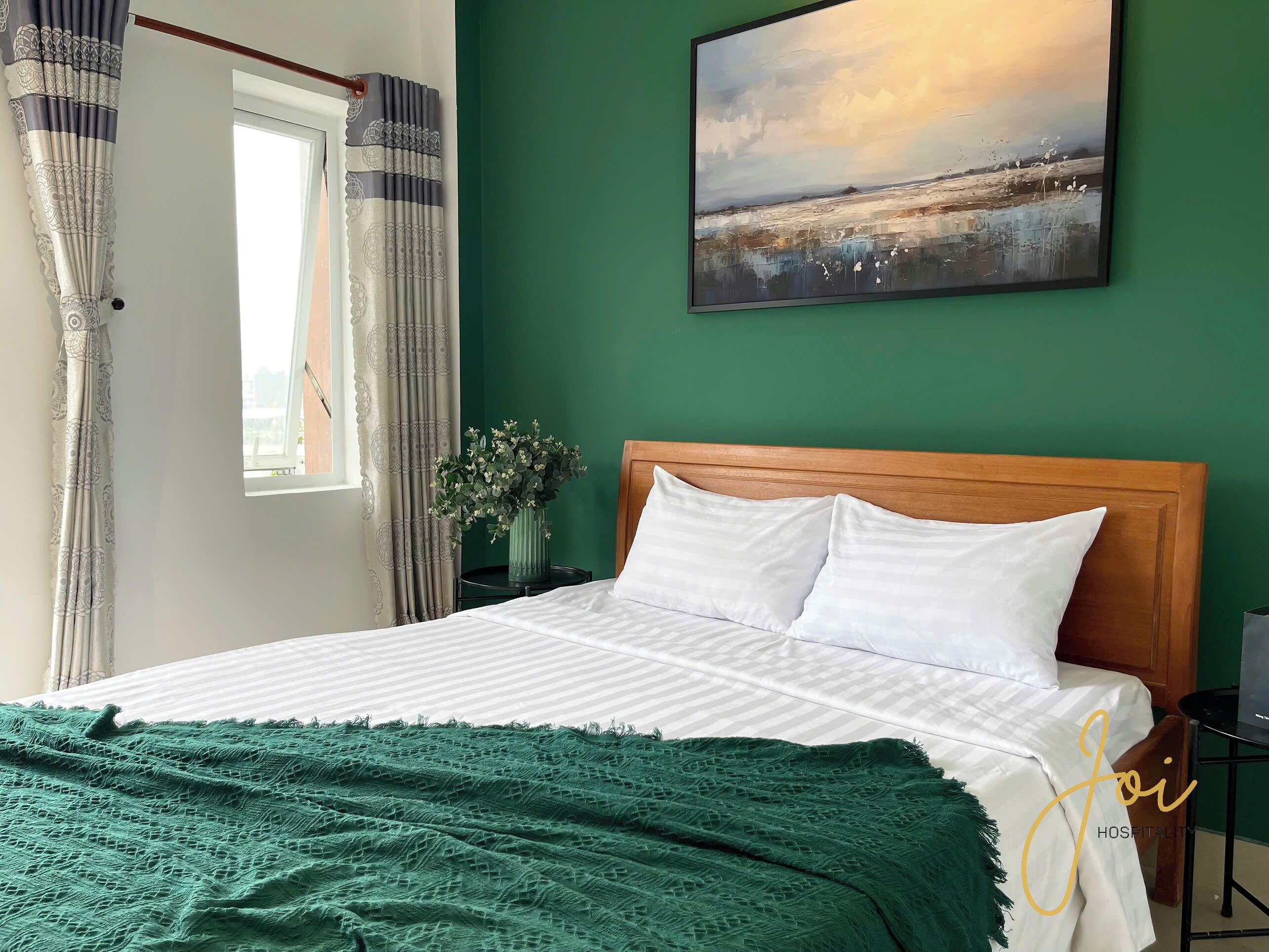 Cama bem arrumada com lençóis brancos e manta verde, cabeceira de madeira e um quadro de paisagem na parede.