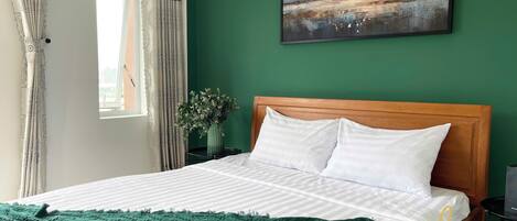 A neatly made bed with white linens and a green throw blanket, a wooden headboard, and a painting of a landscape on the wall.