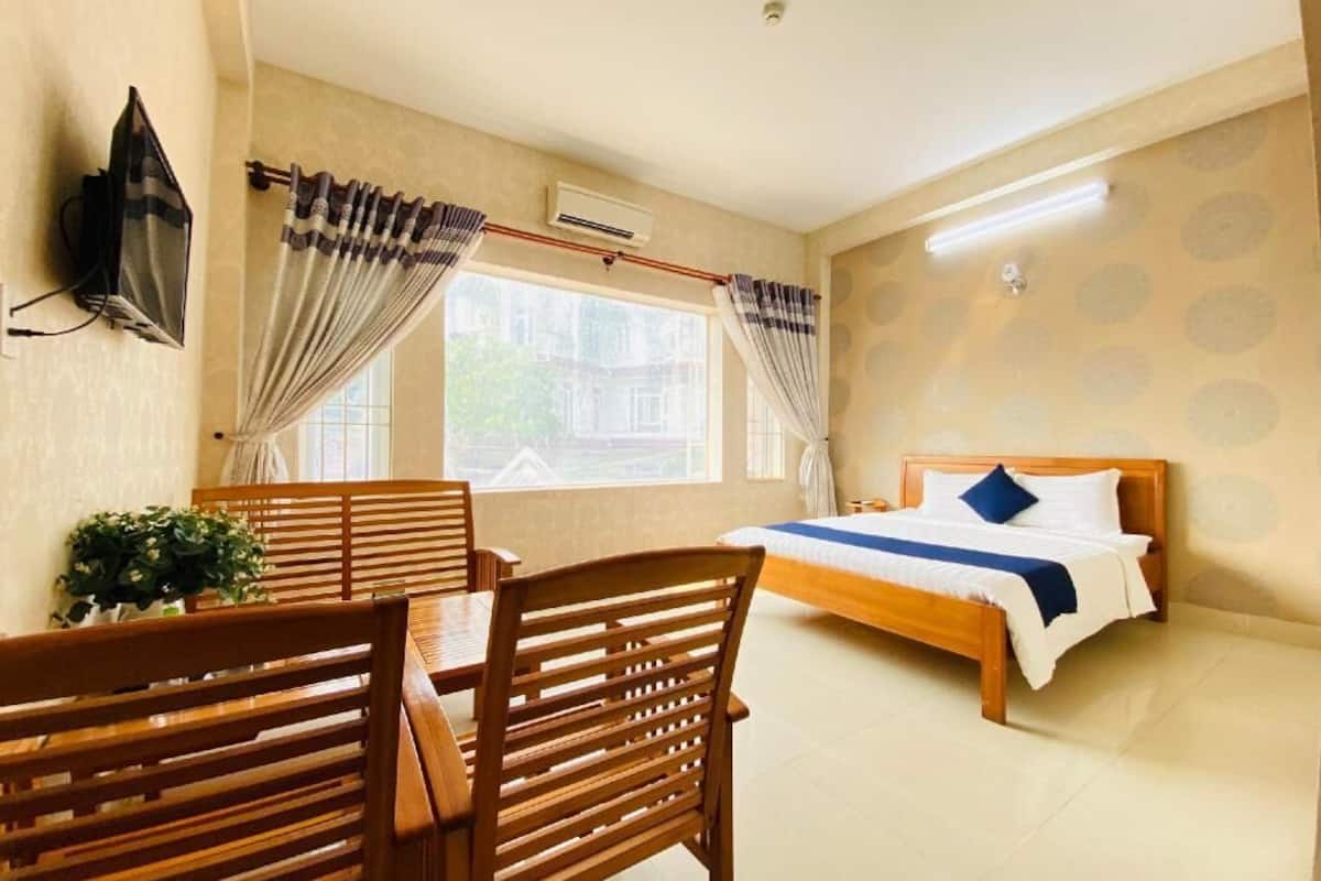 Une chambre d’hôtel avec un lit, une table et des chaises, un téléviseur fixé au mur et une fenêtre avec des rideaux.