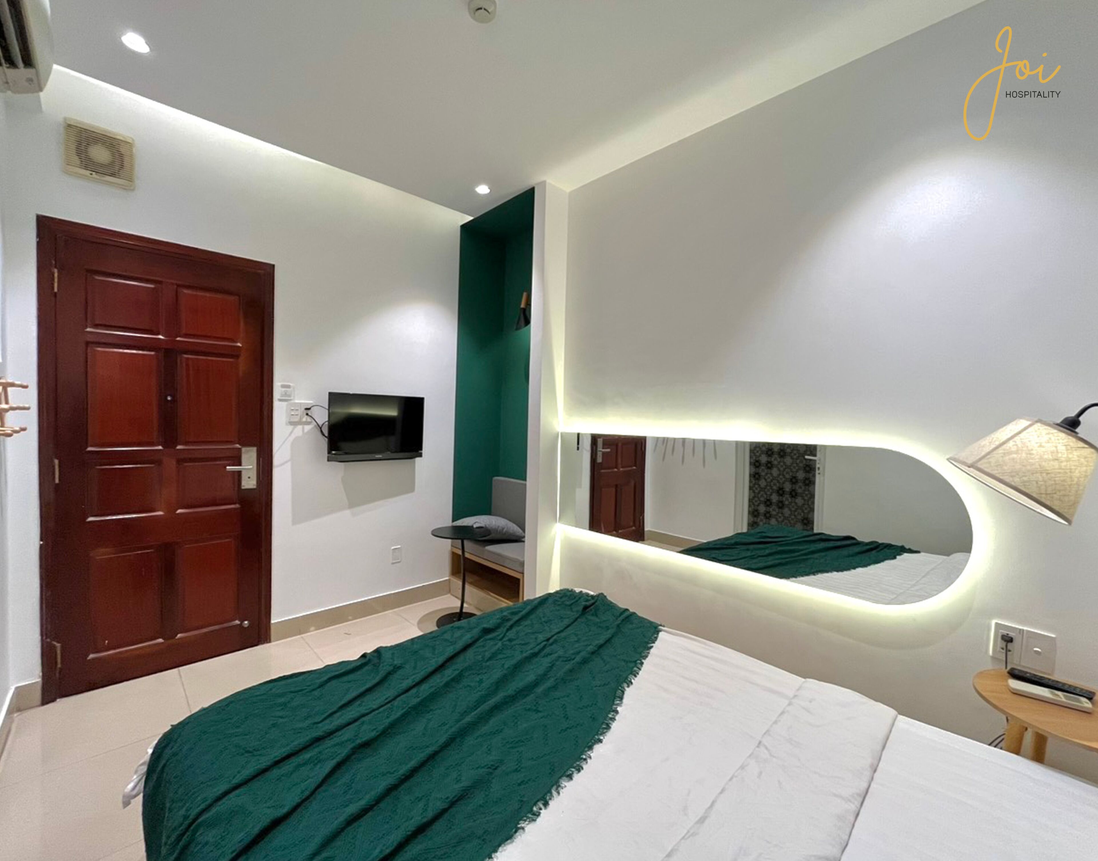 Una habitación de hotel con cama, televisión montada en la pared, espejo y lámpara.