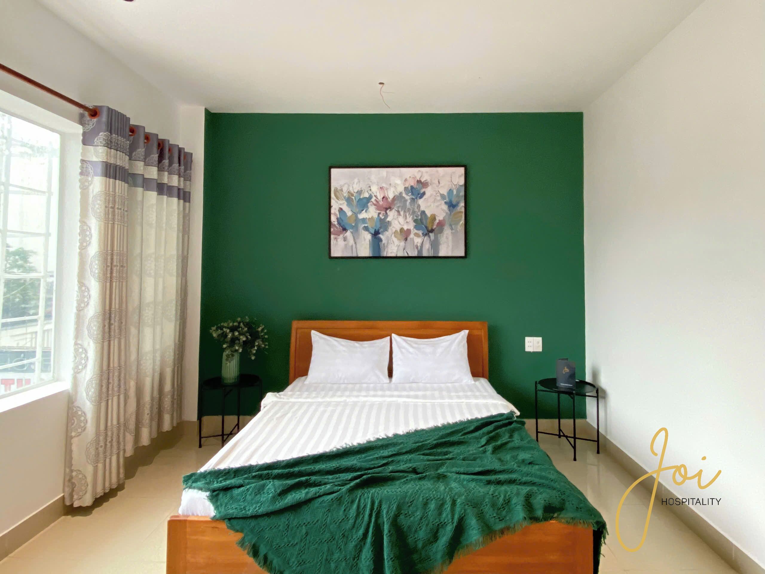 Un dormitorio con pared verde, una cama con ropa de cama blanca, un plaid verde, un cuadro y una ventana con cortinas.