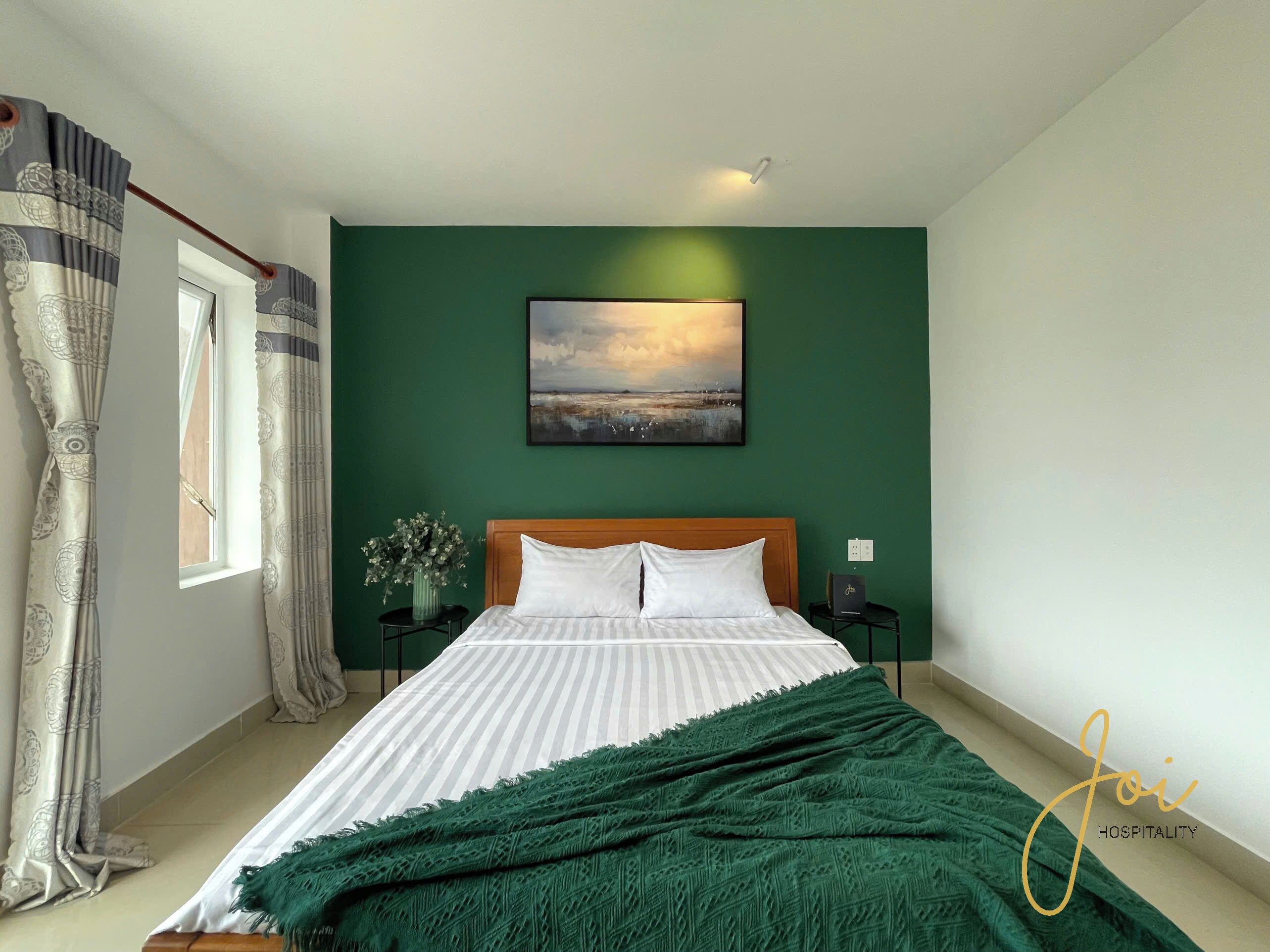 A bedroom with a bed, a nightstand, a vase with flowers, a painting on the wall, and a window with curtains.