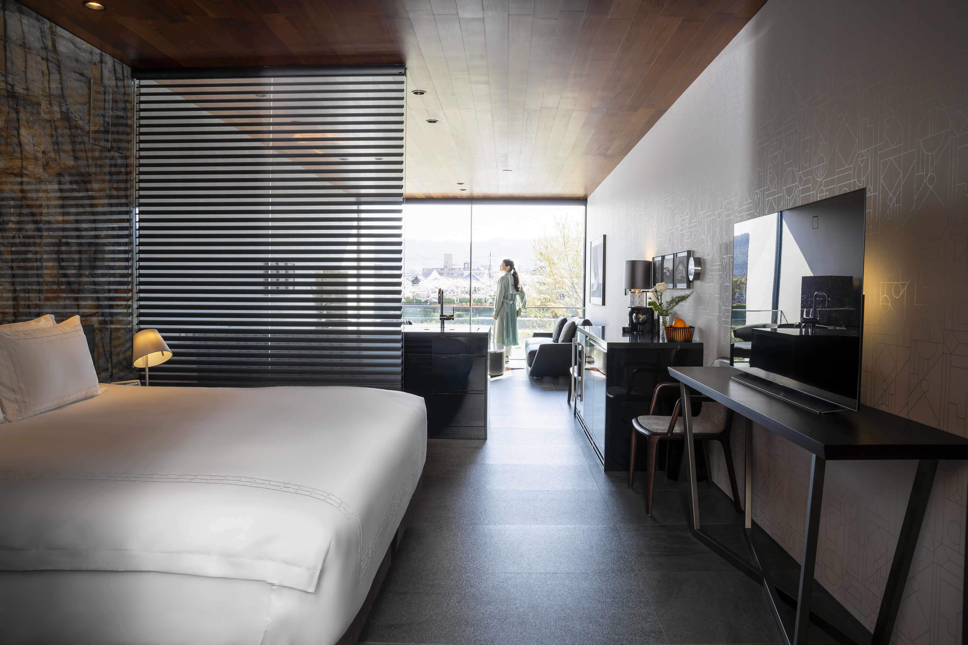A modern hotel room with a large bed, a desk with a flat-screen TV, and a view of the city through large windows.