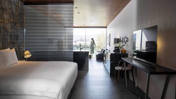 A modern hotel room with a large bed, a desk with a flat-screen TV, and a view of the city through large windows.
