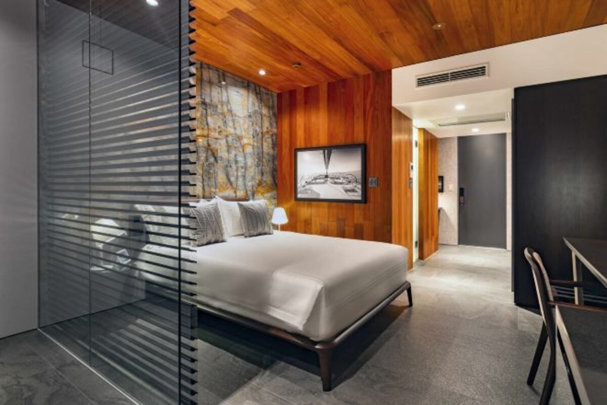 Quarto moderno com cama, divisória de vidro, teto de madeira e um quadro na parede.