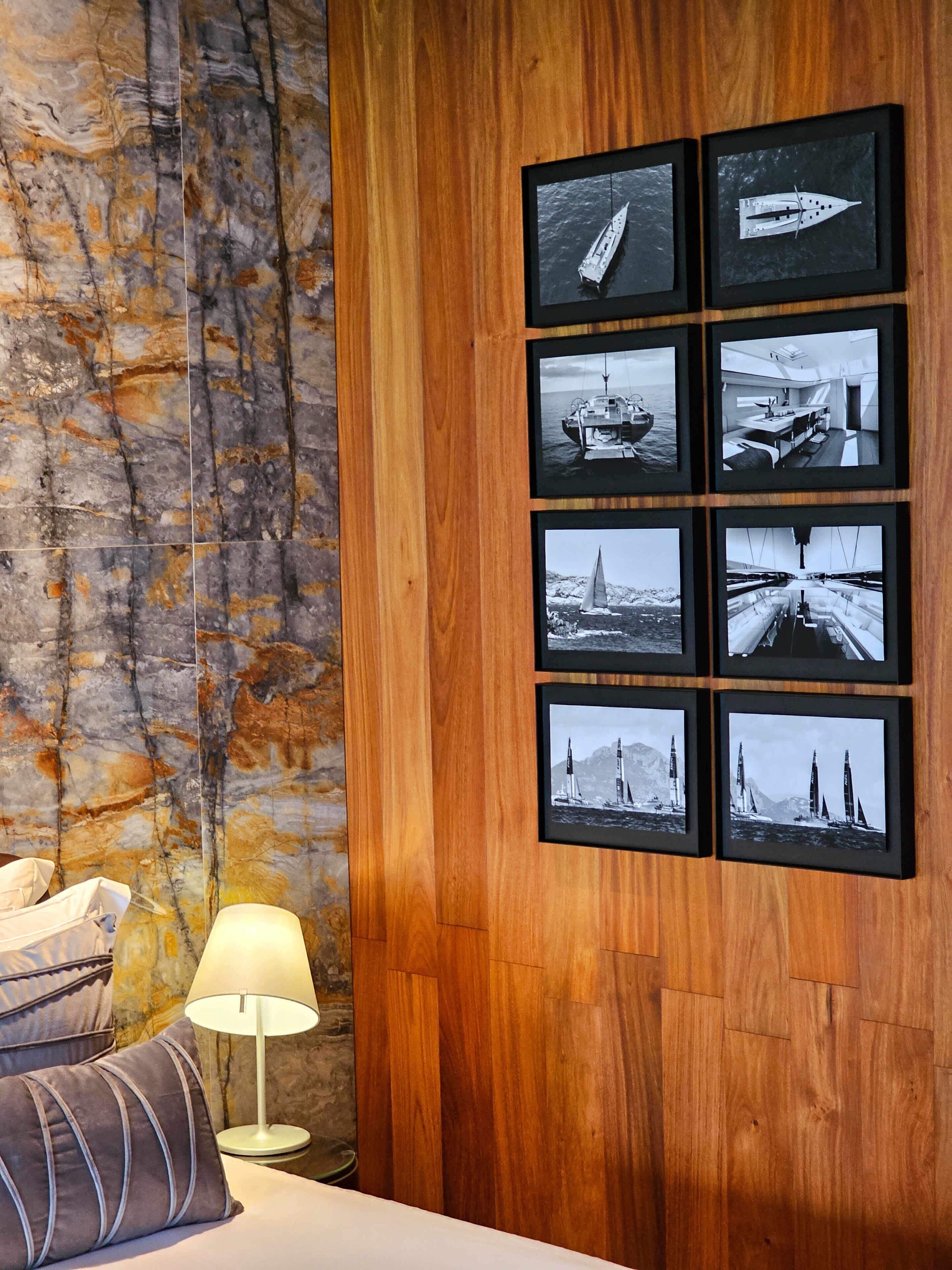 A hotel room with a wooden panel wall, a bed with pillows, a nightstand with a lamp, and a series of framed black-and-white photographs of sailing ships and yachts.