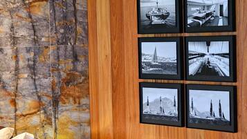 A hotel room with a wooden panel wall, a bed with pillows, a nightstand with a lamp, and a series of framed black-and-white photographs of sailing ships and yachts.