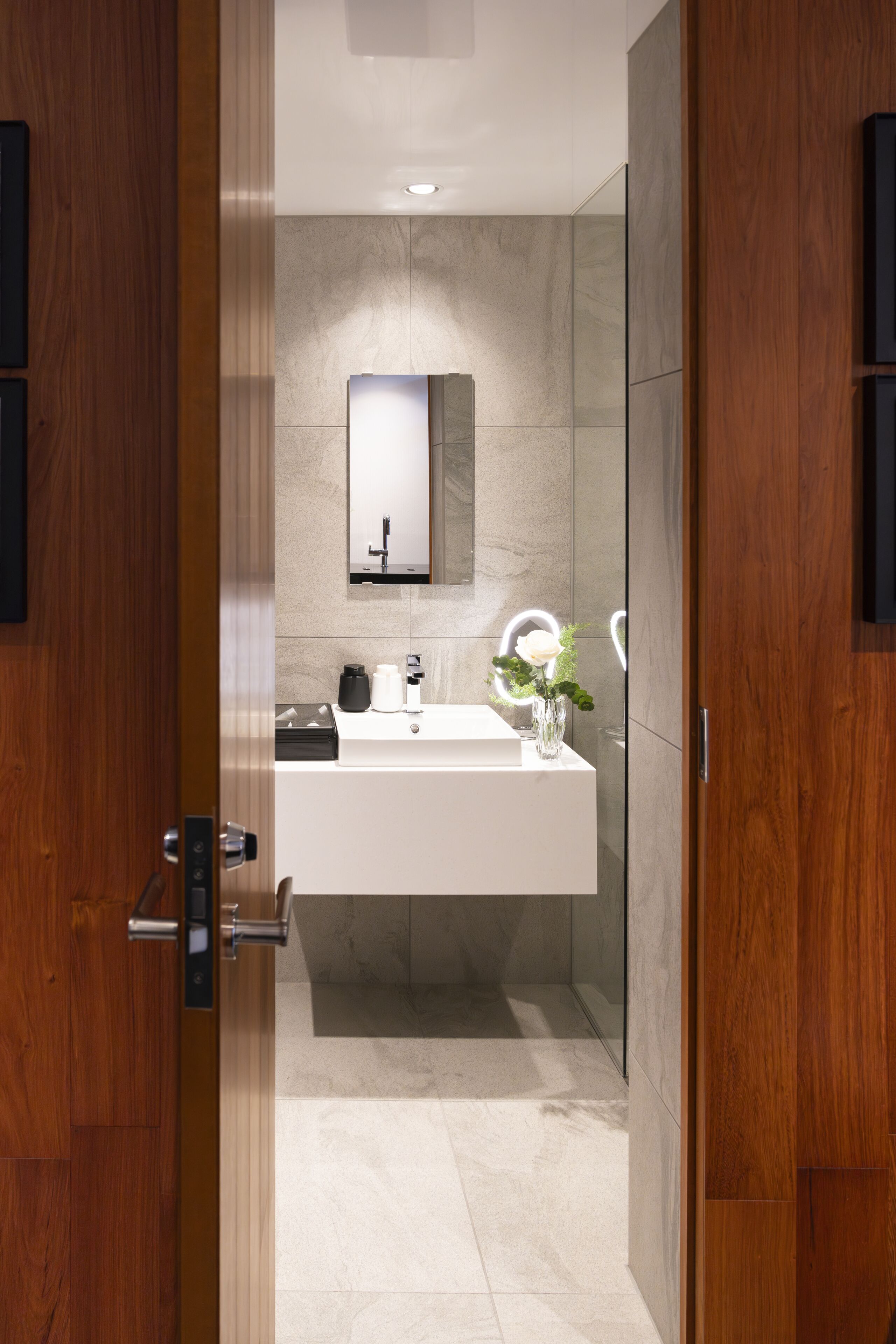 A modern bathroom with a wooden door, a white sink, a mirror, and a vase with flowers.