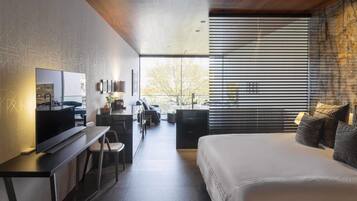 Ein modernes Hotelzimmer mit einem großen Bett, einem Schreibtisch mit Flachbildfernseher, einer Sitzecke mit Sofa und Sesseln sowie einem großen Fenster mit Jalousien.