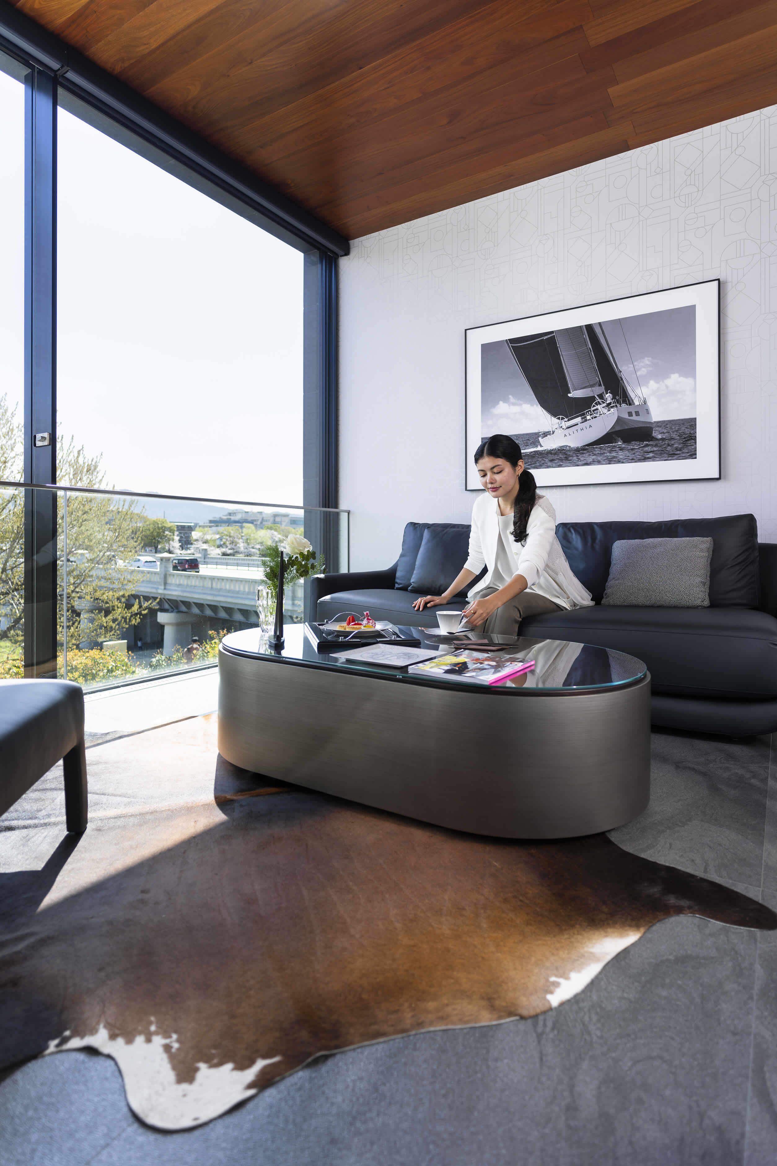 A modern living room with a glass coffee table, a sofa, and a large window offering a view of a bridge and greenery.