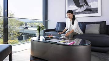 A modern living room with a glass coffee table, a sofa, and a large window offering a view of a bridge and greenery.