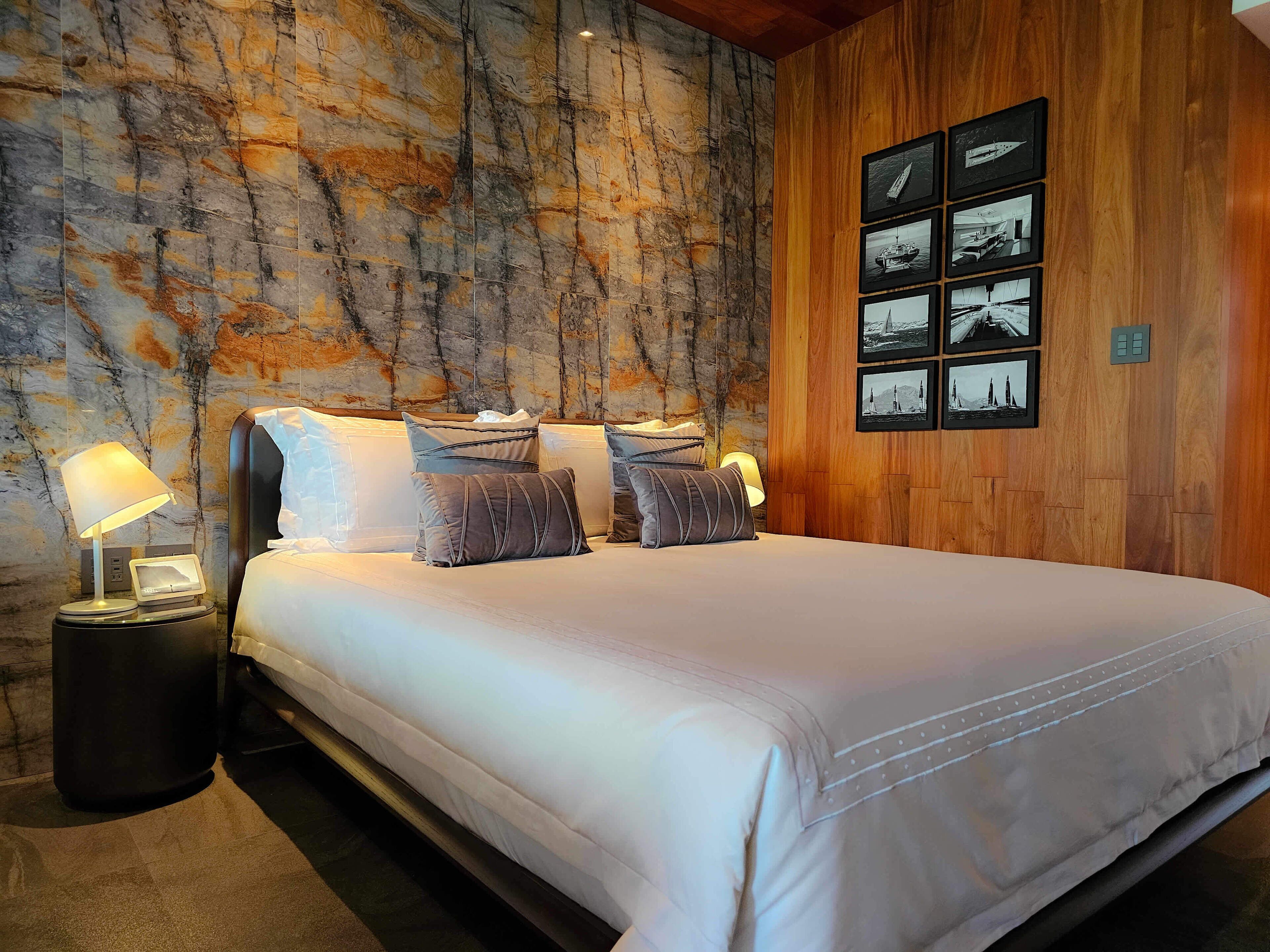 A neatly made bed with a white duvet and multiple pillows, a nightstand with a lamp, and a wall with a wooden panel and framed pictures.