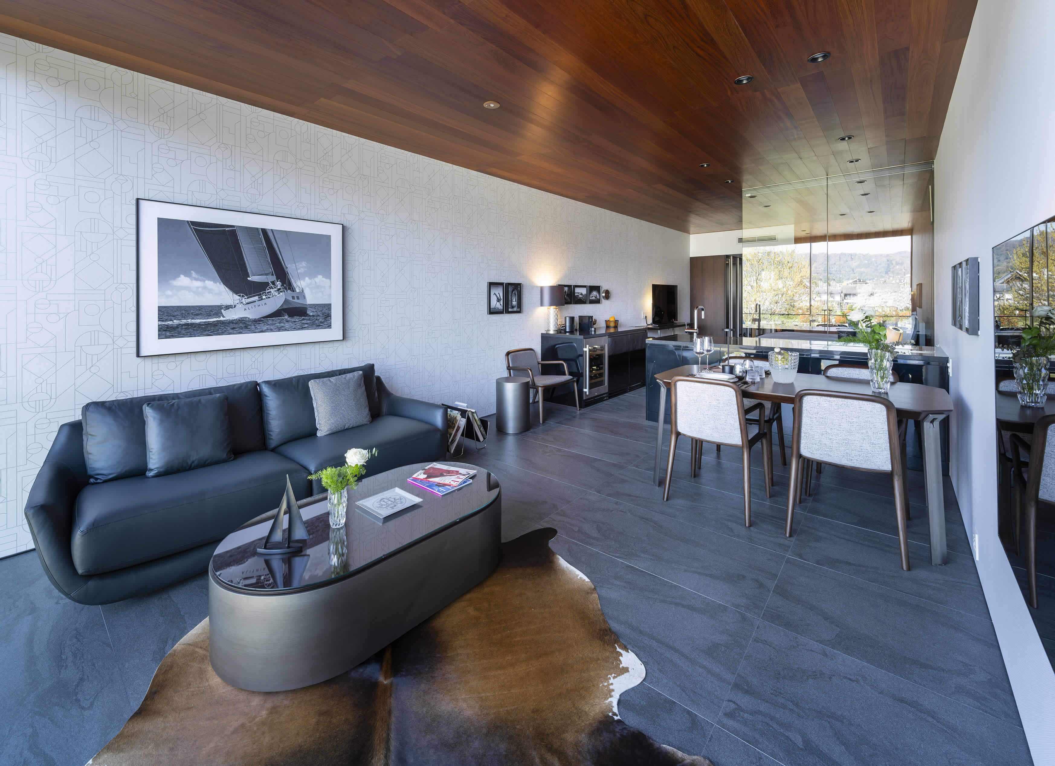 A modern living room with a black leather sofa, a glass coffee table, and a cowhide rug. There is a kitchen area with a dining table and chairs, and a large window offering a view of the outdoors.