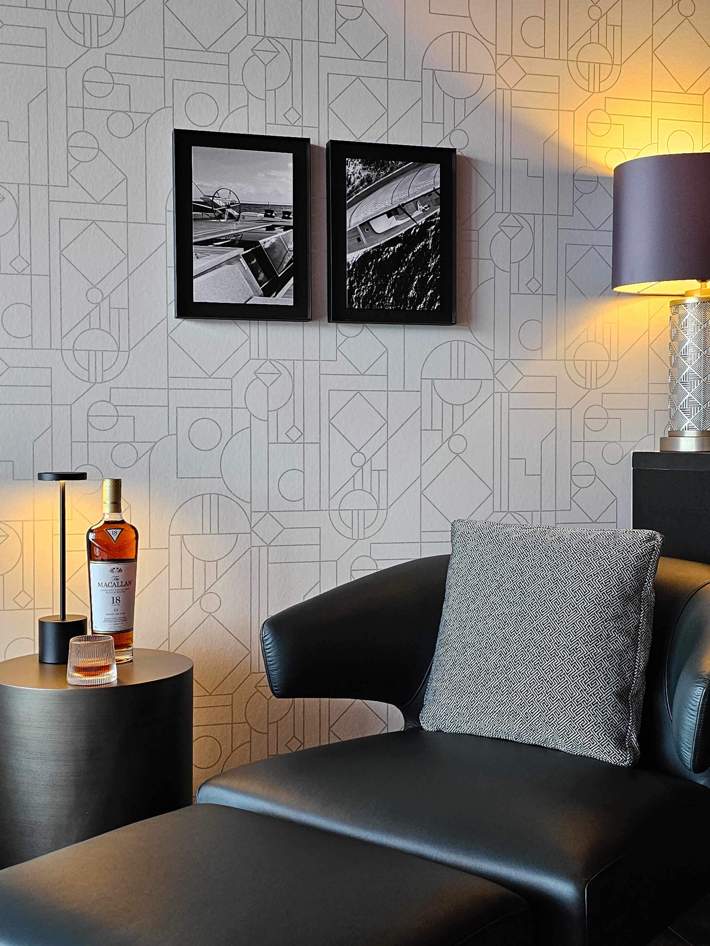 A modern room with a geometric patterned wallpaper, a black leather sofa, a patterned pillow, a side table with a bottle of Macallan whisky, and two framed photographs on the wall.