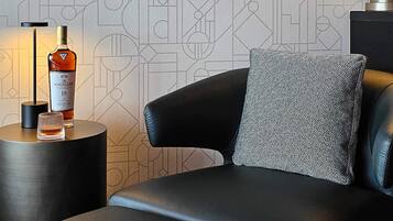 A modern room with a geometric patterned wallpaper, a black leather sofa, a patterned pillow, a side table with a bottle of Macallan whisky, and two framed photographs on the wall.