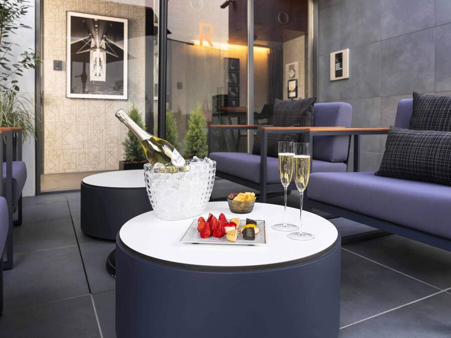 A modern outdoor seating area with a round table, a bottle of champagne, and a plate of fruit.