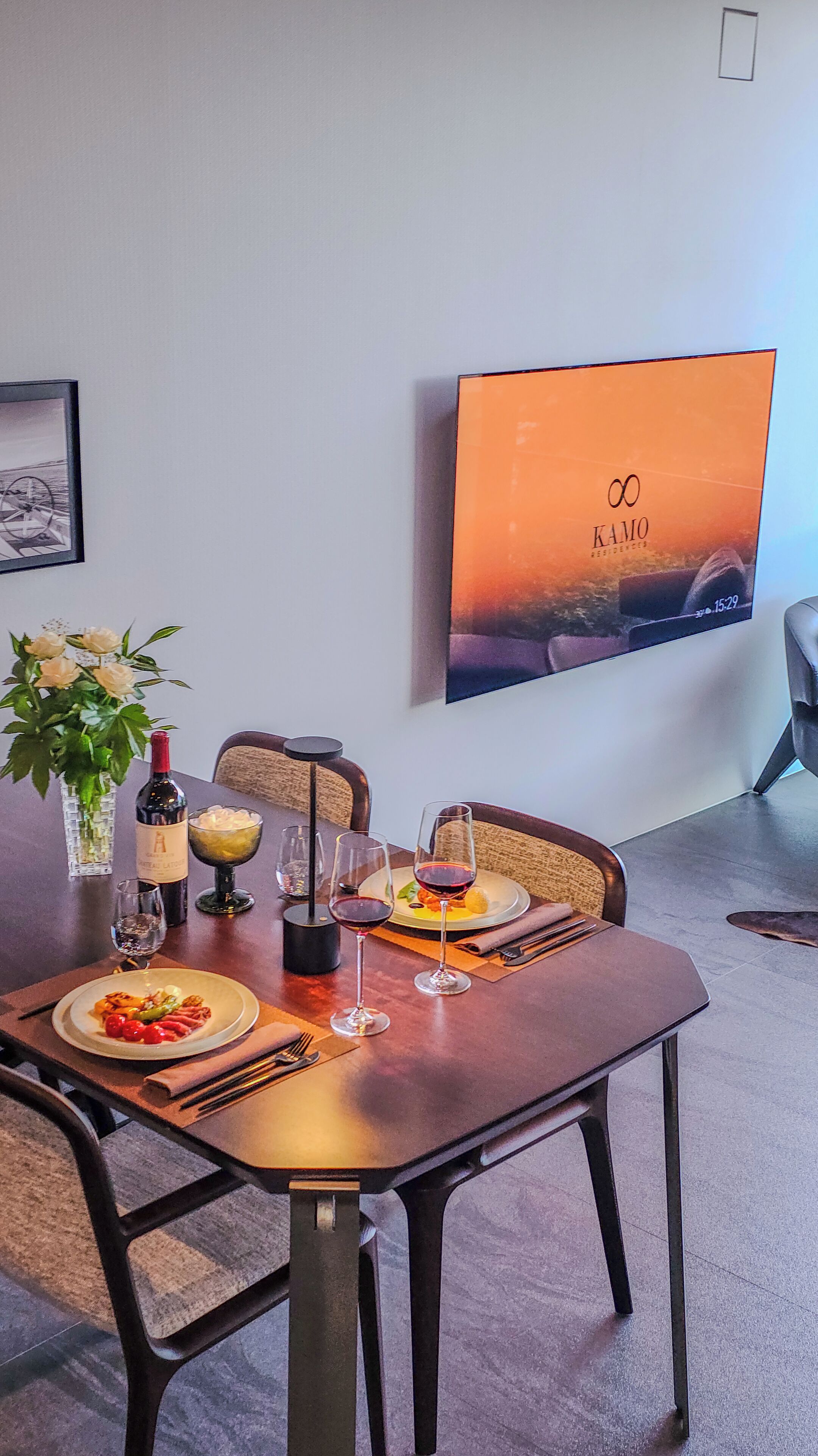 Ein Essbereich mit Tisch für zwei Personen, einer Weinflasche, einer Obstschale und einem Teller mit Essen. An der Wand hängt ein gerahmtes Bild und ein Flachbildfernseher zeigt das Logo 'KAMO RESIDENCES'.