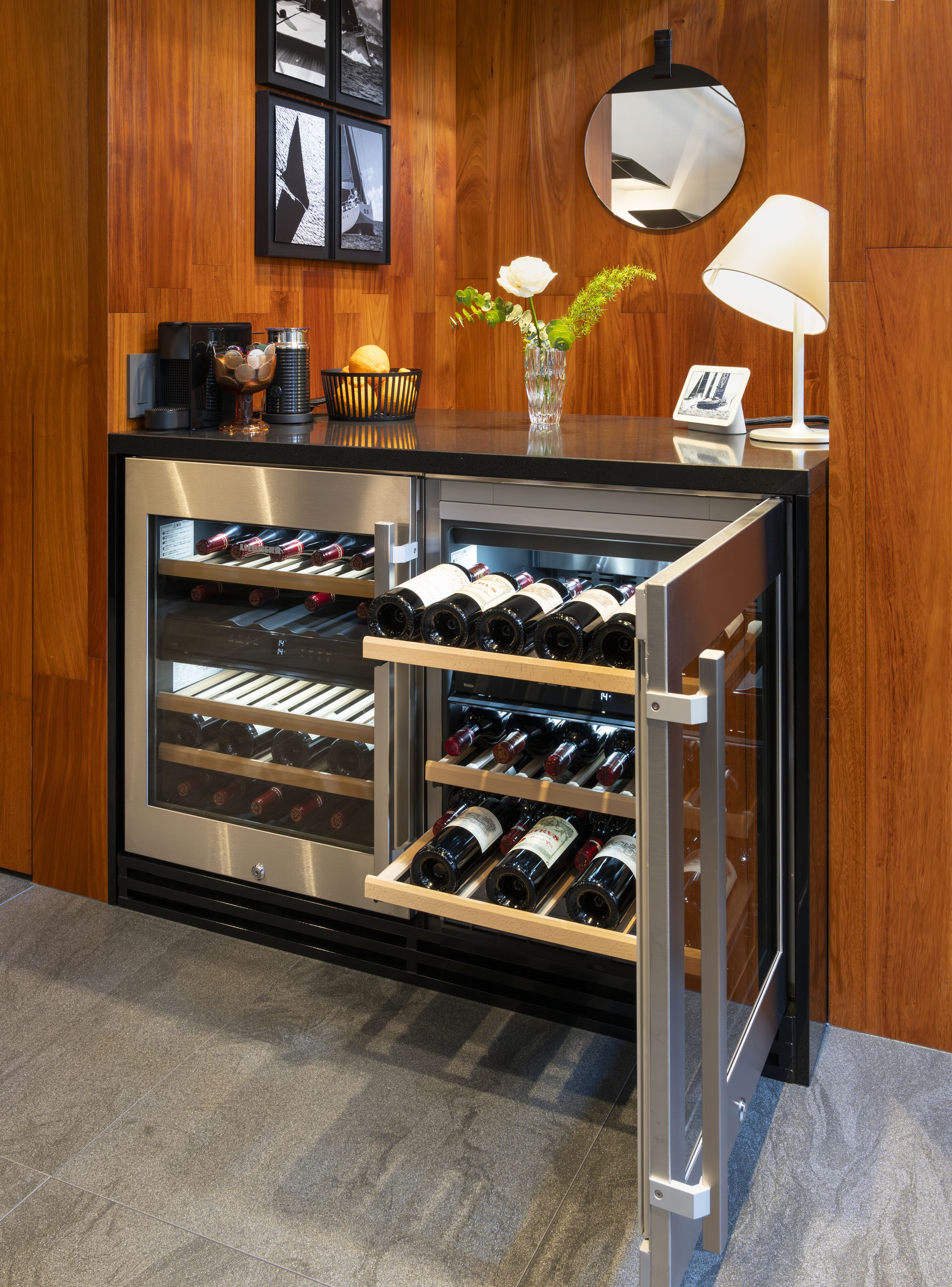 A wine cooler with multiple shelves of wine bottles, a small table with a vase and a lamp, and framed pictures on the wall.