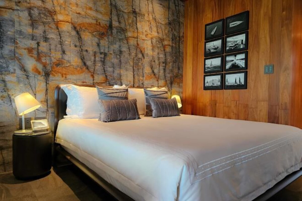 Quarto com cama, duas mesas de cabeceira com luminárias, parede de madeira com textura e uma série de quadros na parede.