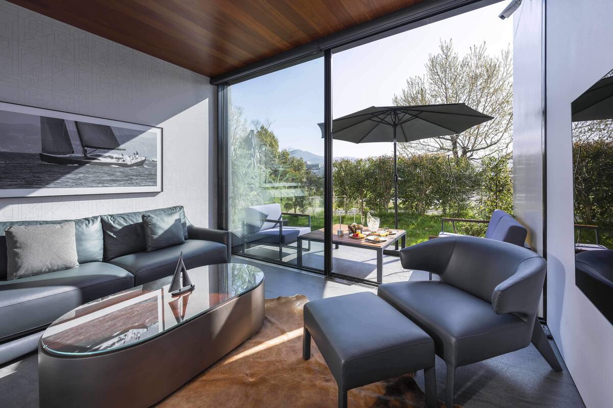 A modern living room with a glass coffee table, leather sofa, and a large window offering a view of greenery and a sailboat.